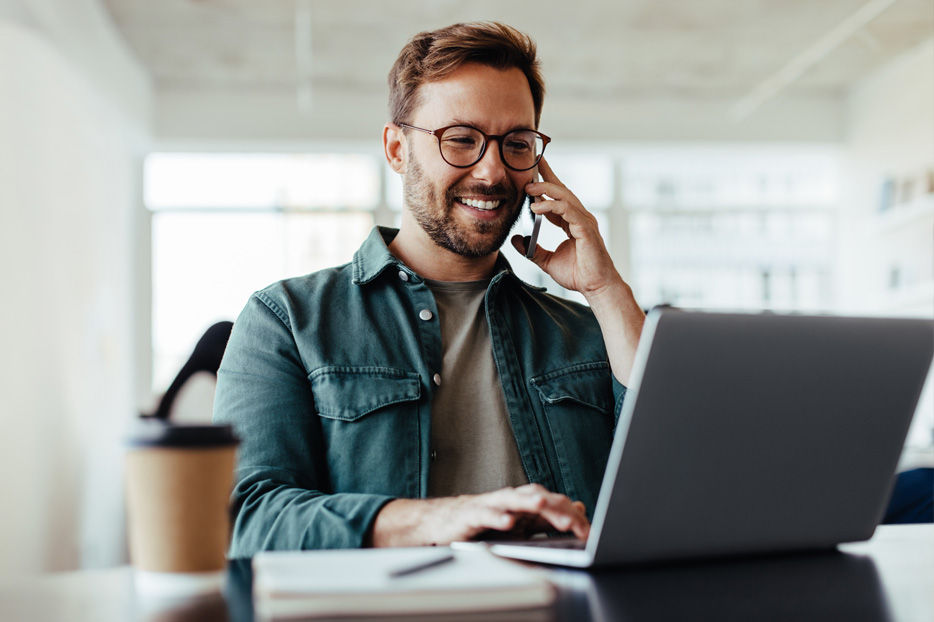 A man with his laptop open. He's holding a mobile phone to his ear.